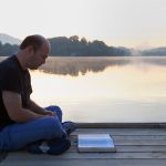 Man reading a book on a wooden bridge surrounded by hills and a lake under the sunlight
